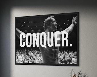 May include: A black and white framed wall art print featuring a muscular bodybuilder posing on stage with arms raised, overlaid with the bold white text CONQUER. The rectangular print is displayed in a thin black frame on a dark grey wall.
