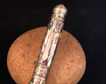 May include: A silver and abalone pen with a detailed design on the cap and barrel. The pen is resting on a brown rock.