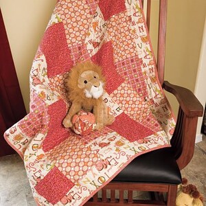 May include: A red and white patchwork quilt with a lion stuffed animal on top. The quilt has a white border with letters printed on it. The quilt is on a wooden rocking chair.