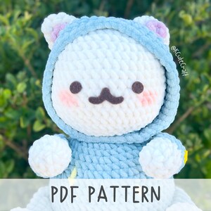 Op de afbeelding: Een wit gebreid pluche speelgoed dat een blauwe hoodie met roze oren draagt. Het speelgoed heeft een schattig cartoon gezicht met bruine ogen, een bruine snor en roze wangen. De tekst "PDF PATTERN" staat onderaan de afbeelding.