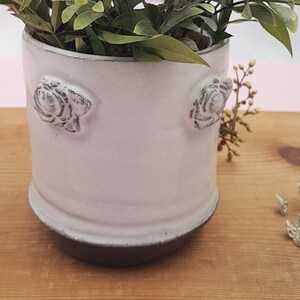 May include: White ceramic planter with a textured rose design on the sides. The planter has a dark gray base and is filled with green leafy plants.