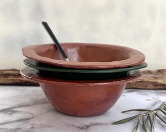 May include: A set of three ceramic bowls, one red, one green, and one brown. The bowls are stacked on top of each other. The red bowl is the largest and the brown bowl is the smallest.