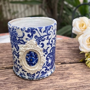 May include: A white ceramic planter with a blue and white floral pattern. The planter has a decorative silver frame with a blue floral design in the center.