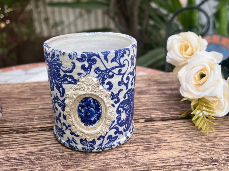 May include: A white ceramic planter with a blue and white floral pattern. The planter has a decorative silver frame with a blue floral design in the center.