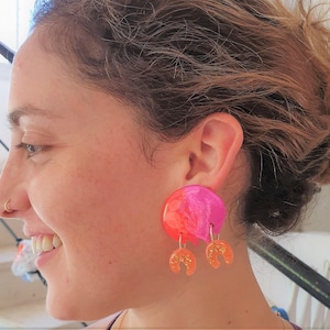 Könnte beinhalten: Auffällige Ohrringe in Pink, Orange und Gold. Die Ohrringe haben einen kreisförmigen oberen Teil mit Marmoreffekt und zwei hängende, halbmondförmige Anhänger mit Glitzer. Ein auffälliges Schmuckstück.