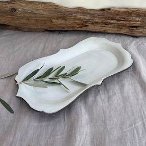 May include: White ceramic serving tray with a scalloped edge and a dark gray rim. The tray is empty and has a sprig of green leaves on top.