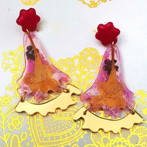 以下が含まれることがあります： ゴールドのアクセントが付いたピンクのレジン製のイヤリングのペア。各イヤリングには、レジンに閉じ込められたドライフラワーと星型の赤いトップが付いています。