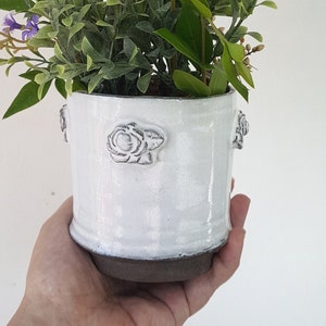 May include: White ceramic planter with raised rose details, holding a small arrangement of purple flowers and green foliage.