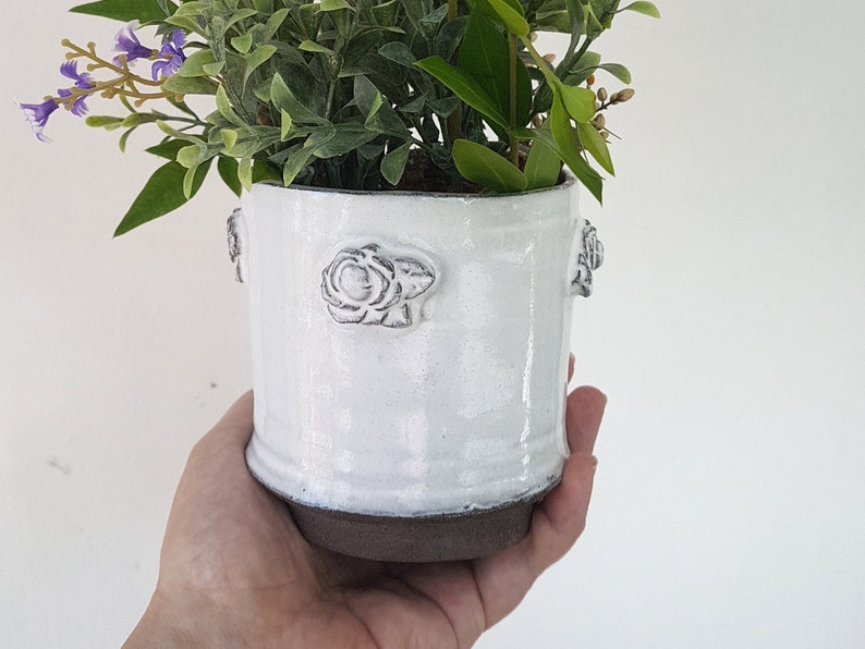 May include: White ceramic planter with raised rose details, holding a small arrangement of purple flowers and green foliage.
