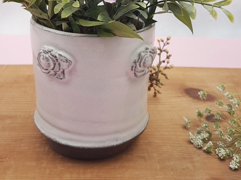 May include: White ceramic planter with a textured rose design on the sides. The planter has a dark gray base and is filled with green leafy plants.