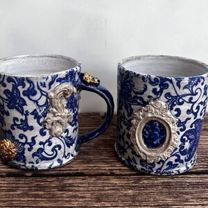 May include: Two blue and white ceramic mugs with gold accents. The mugs have a floral pattern and ornate details.
