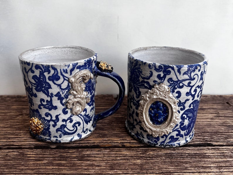 May include: Two blue and white ceramic mugs with gold accents. The mugs have a floral pattern and ornate details.