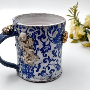May include: A white ceramic mug with a blue and white floral pattern. The mug has a blue handle and gold accents.