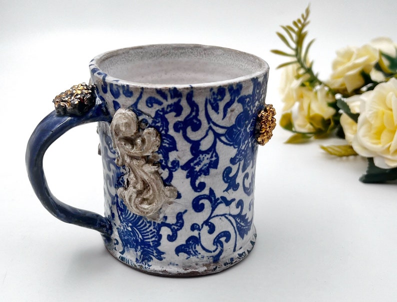 May include: A white ceramic mug with a blue and white floral pattern. The mug has a blue handle and gold accents.