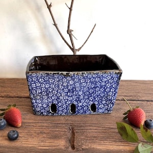 Op de afbeelding: Een rechthoekige keramische bessenmand met een blauw-wit bloemenpatroon aan de buitenkant en een donkerbruine binnenkant. De onderkant van de mand heeft drie ronde afvoergaten aan de voorzijde, met een glanzende afwerking.