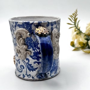 May include: A white ceramic mug with a blue and white floral pattern, a blue handle, and gold accents.