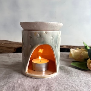 May include: A ceramic oil burner with a moon and star design. The burner has a small dish on top for essential oils and a space for a tea light candle underneath. The burner is lit and the candle is glowing.
