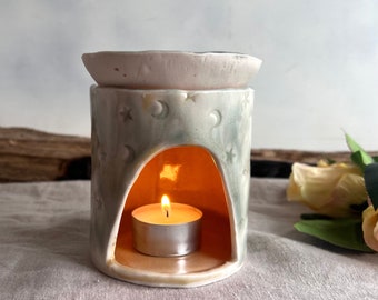 May include: A cylindrical ceramic wax warmer featuring a light green and white marbled glaze with embossed star and moon motifs. The piece has a wide arched opening at the front containing a lit tea light candle and a shallow dish on top.