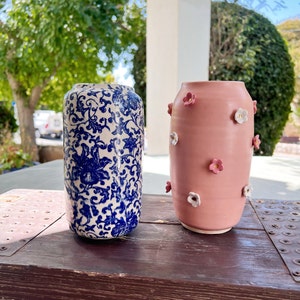 Könnte beinhalten: Zwei zylindrische Keramikvasen. Eine Vase ist mit einem komplexen blau-weißen Blumenmuster über die gesamte Oberfläche versehen. Die zweite Vase ist einfarbig mattrosa und mit kleinen, erhabenen weißen und rosa Blumenapplikationen verziert.