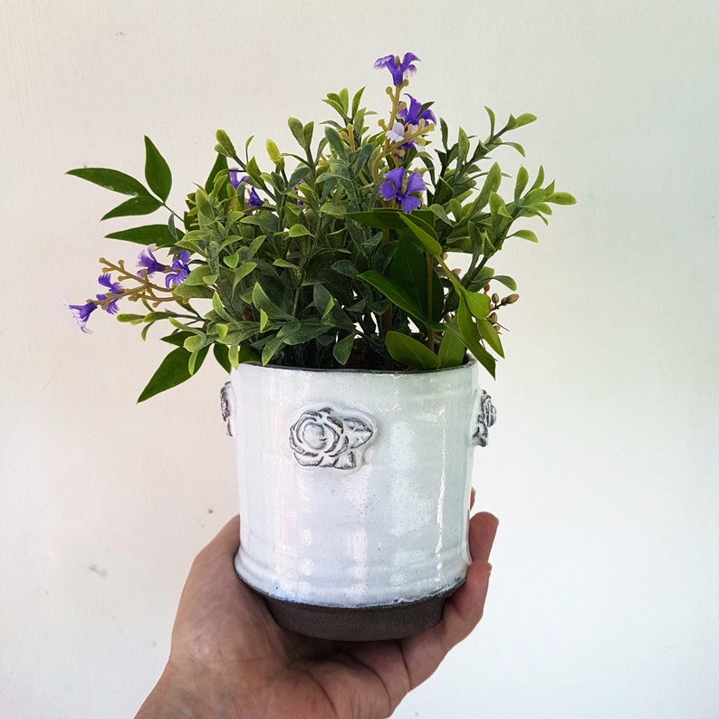 May include: A white ceramic planter with a raised floral design, filled with a small green plant with purple flowers.