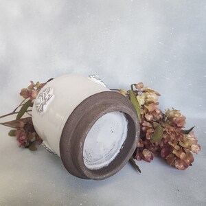 May include: White ceramic vase with a brown rim and embossed floral details. The vase is sitting on a light gray surface with dried flowers.