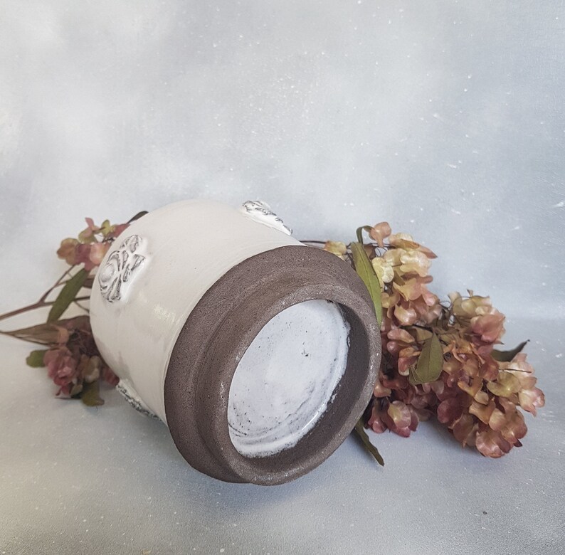 May include: White ceramic vase with a brown rim and embossed floral details. The vase is sitting on a light gray surface with dried flowers.