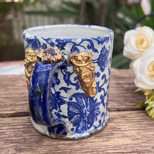 May include: A small blue and white ceramic mug with a gold floral design and gold accents on the handle. The mug is sitting on a wooden surface.