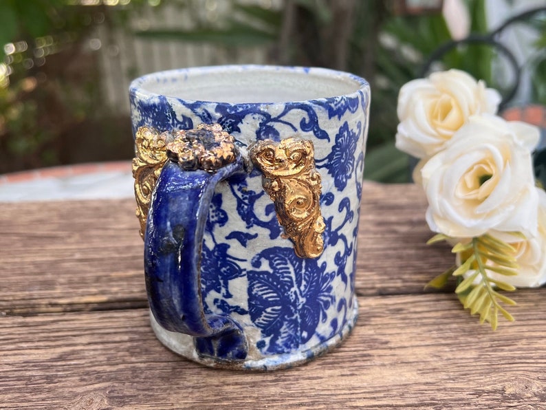 May include: A small blue and white ceramic mug with a gold floral design and gold accents on the handle. The mug is sitting on a wooden surface.