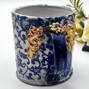 May include: A white ceramic mug with a blue and gold floral pattern. The mug has a blue handle with gold accents.