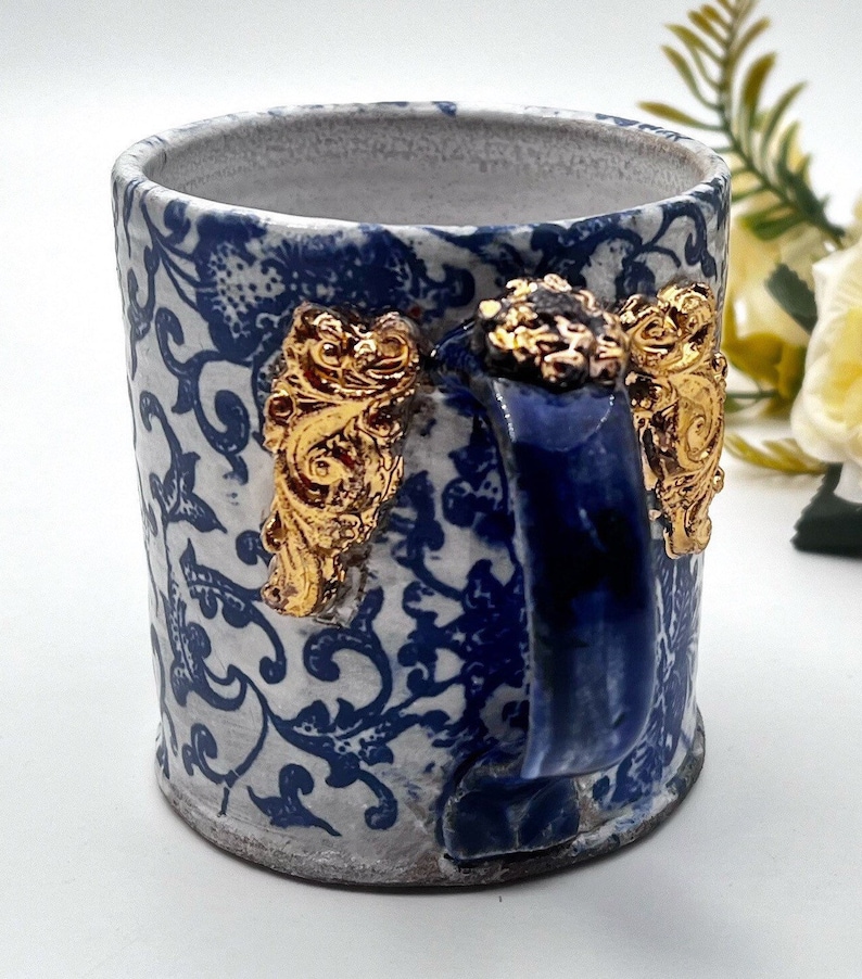 May include: A white ceramic mug with a blue and gold floral pattern. The mug has a blue handle with gold accents.