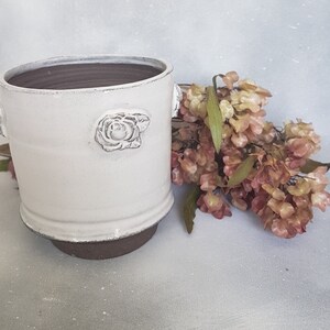 May include: White ceramic planter with a raised rose design and brown rim. The planter is sitting on a white surface with a bouquet of dried flowers.