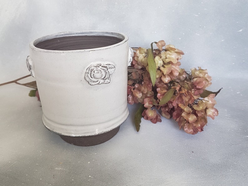 May include: White ceramic planter with a raised rose design and brown rim. The planter is sitting on a white surface with a bouquet of dried flowers.