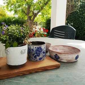 May include: Three ceramic planters with floral designs. The planters are white with blue floral patterns. The planters are sitting on a wooden surface.