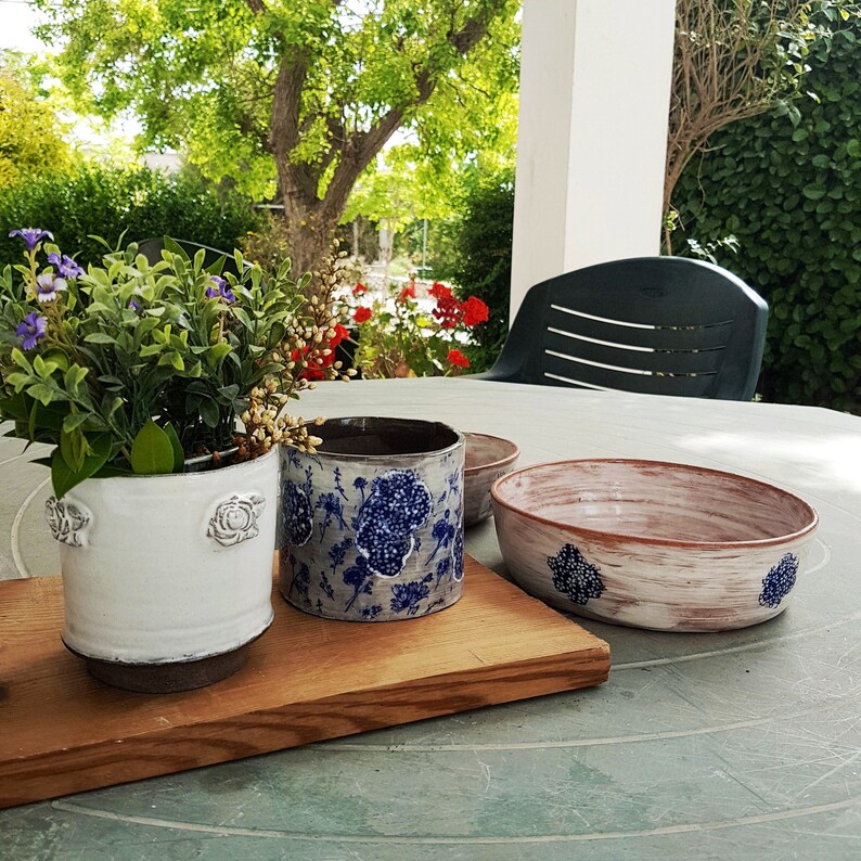 May include: Three ceramic planters with floral designs. The planters are white with blue floral patterns. The planters are sitting on a wooden surface.