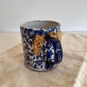May include: A white ceramic mug with a blue and white floral pattern and gold accents on the handle. The mug has a blue handle.