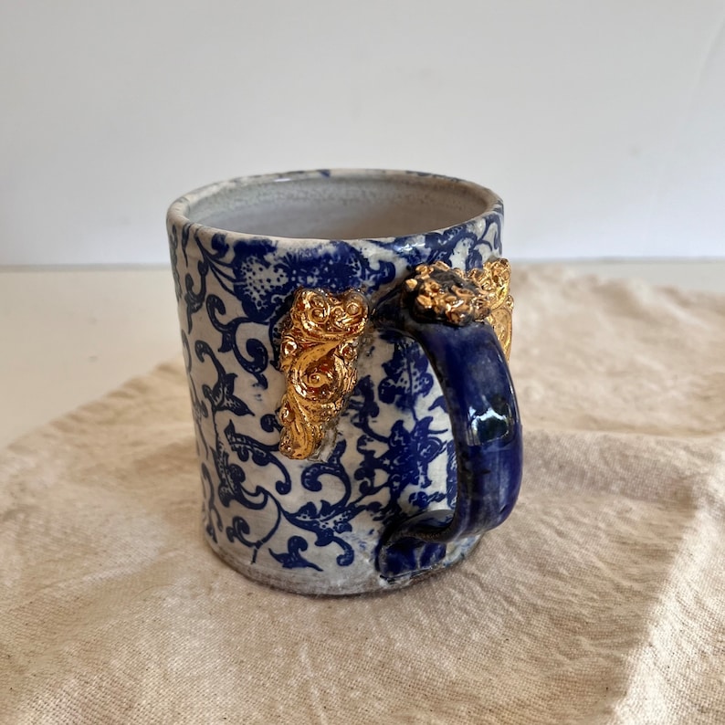 May include: A white ceramic mug with a blue and white floral pattern and gold accents on the handle. The mug has a blue handle.
