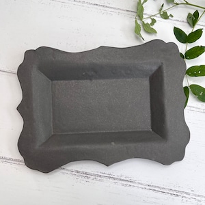 May include: A dark grey ceramic tray with a decorative, scalloped edge. The tray has a rectangular shape with a raised rim.