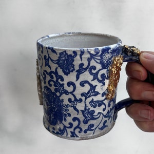 May include: A white ceramic mug with a blue and white floral pattern and gold accents on the handle. The mug is being held by a hand.