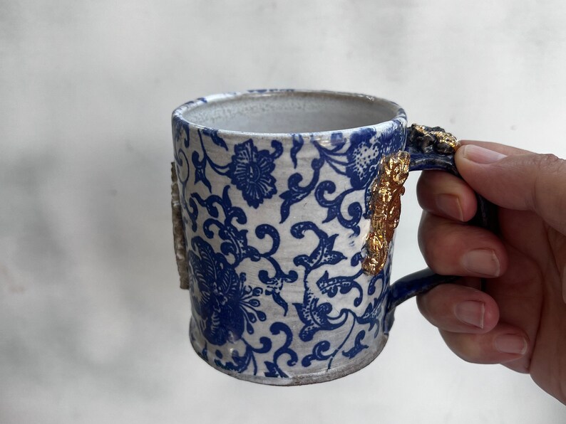 May include: A white ceramic mug with a blue and white floral pattern and gold accents on the handle. The mug is being held by a hand.