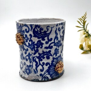 May include: White ceramic cup with a blue and white floral pattern and gold accents. The cup is approximately 4 inches tall.