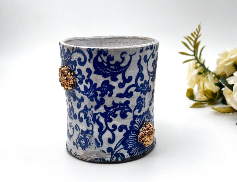 May include: White ceramic cup with a blue and white floral pattern and gold accents. The cup is approximately 4 inches tall.