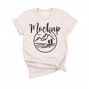 May include: A beige t-shirt with a black graphic of mountains, trees, and a lake. The text "Mockup" is written in black cursive above the graphic.