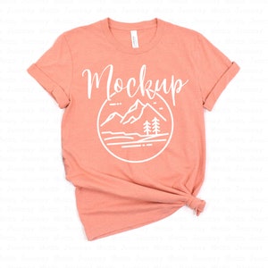 May include: A coral colored t-shirt with a white graphic of mountains, trees, and a lake in a circle. The text "Mockup" is written in a cursive font above the graphic.