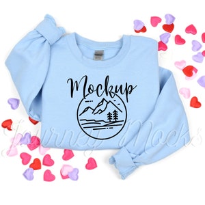May include: A light blue sweatshirt with a black mountain graphic and the word "Mockup" printed on the front. The sweatshirt is surrounded by pink and purple hearts.