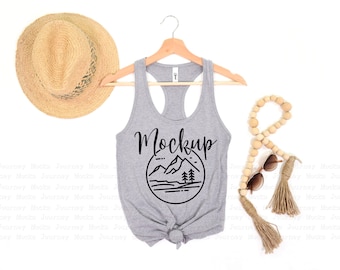May include: A heather gray tank top with the word "Mockup" and a mountain graphic. A straw hat and a beaded garland with sunglasses are also in the image. The tank top is hanging on a wooden hanger.