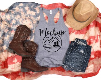 May include: Gray tank top with a black mountain graphic and the word "Mockup" in cursive. The tank top is laying on a red, white, and blue American flag blanket with a pair of brown cowboy boots, a straw cowboy hat, and blue denim shorts.