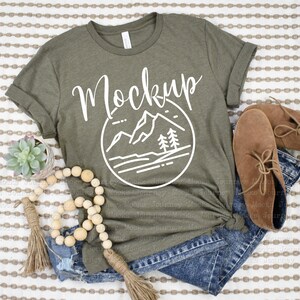 May include: A green t-shirt with a white graphic of mountains and trees in a circle. The text "Mockup" is written above the graphic. The t-shirt is laying on a striped fabric with a pair of brown boots, a wooden bead garland, and a small potted succulent.