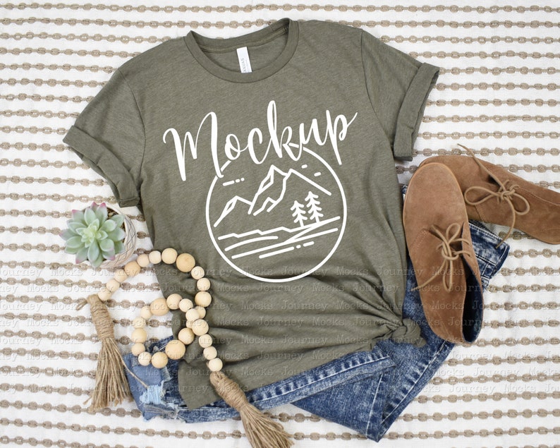 May include: A green t-shirt with a white graphic of mountains and trees in a circle. The text "Mockup" is written above the graphic. The t-shirt is laying on a striped fabric with a pair of brown boots, a wooden bead garland, and a small potted succulent.