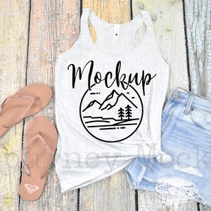 May include: A white tank top with a black mountain range graphic and the word "Mockup" in black script. The tank top is laying on a wooden surface with a pair of brown flip flops and a pair of light blue denim shorts with white lace trim.