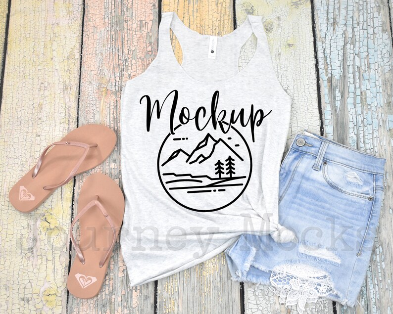 May include: A white tank top with a black mountain range graphic and the word "Mockup" in black script. The tank top is laying on a wooden surface with a pair of brown flip flops and a pair of light blue denim shorts with white lace trim.
